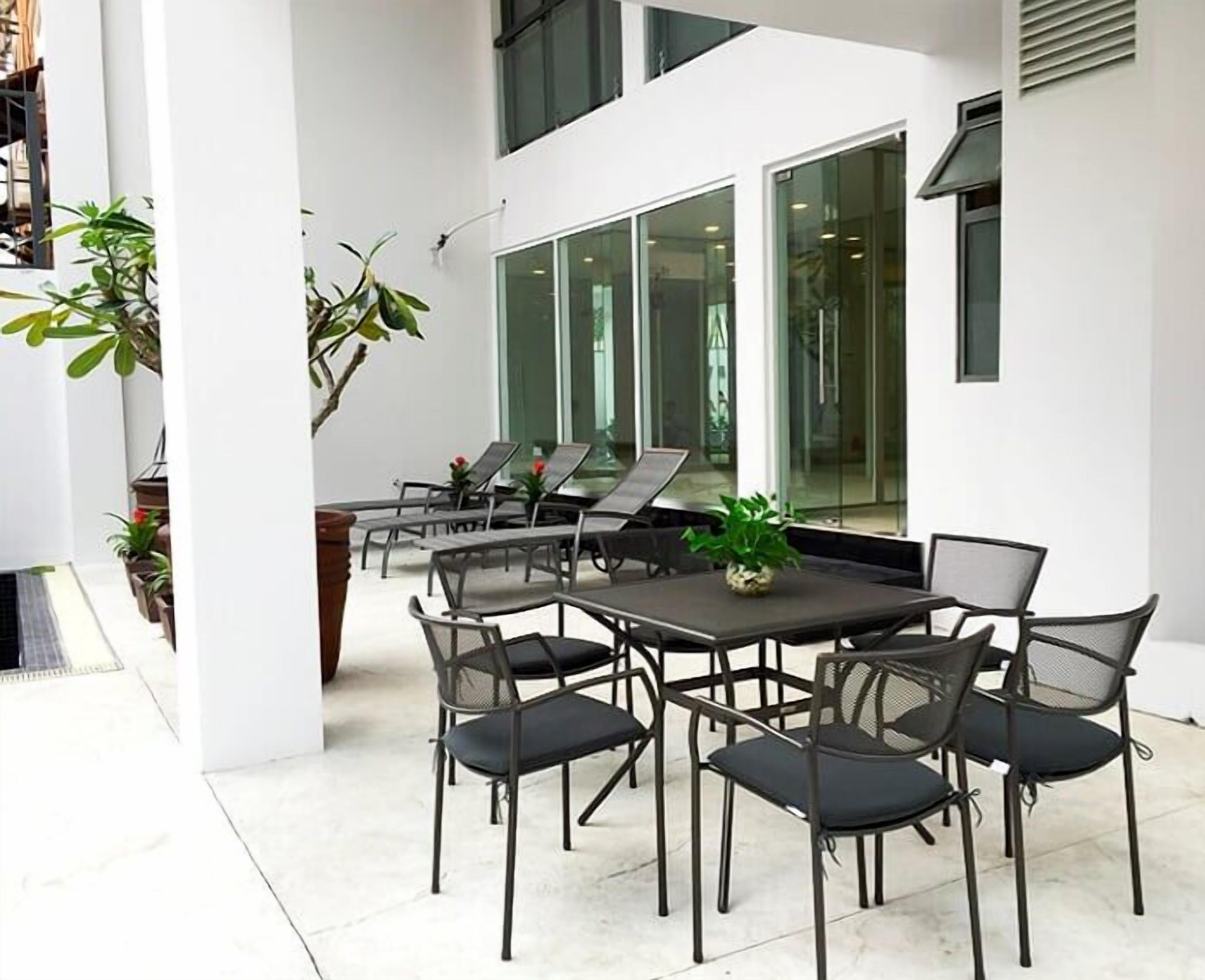 A modern outdoor patio with a table and chairs, surrounded by potted plants and large windows.