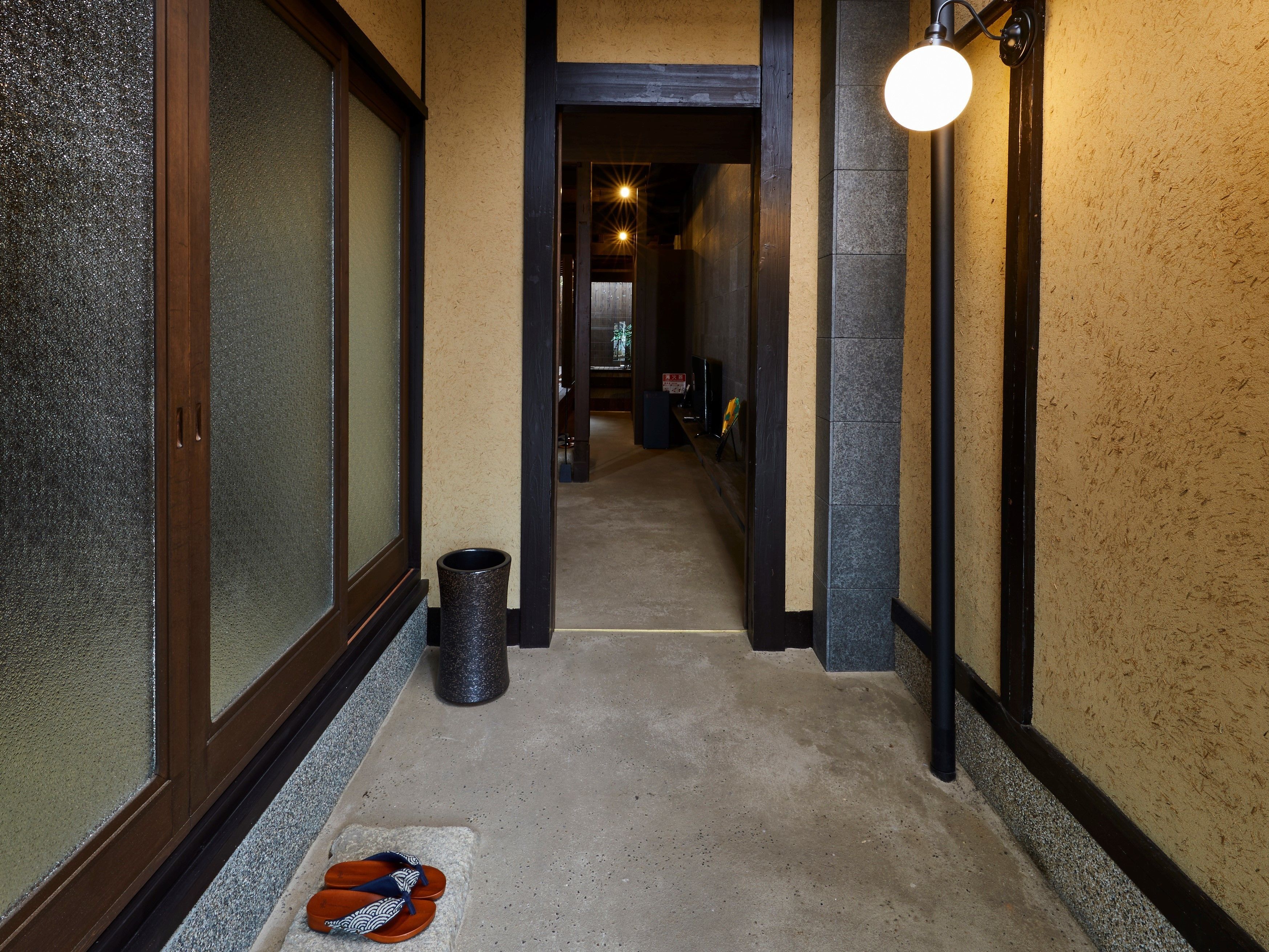 A hallway with a trash bin, a pair of flip-flops on the floor, and a light fixture on the wall.