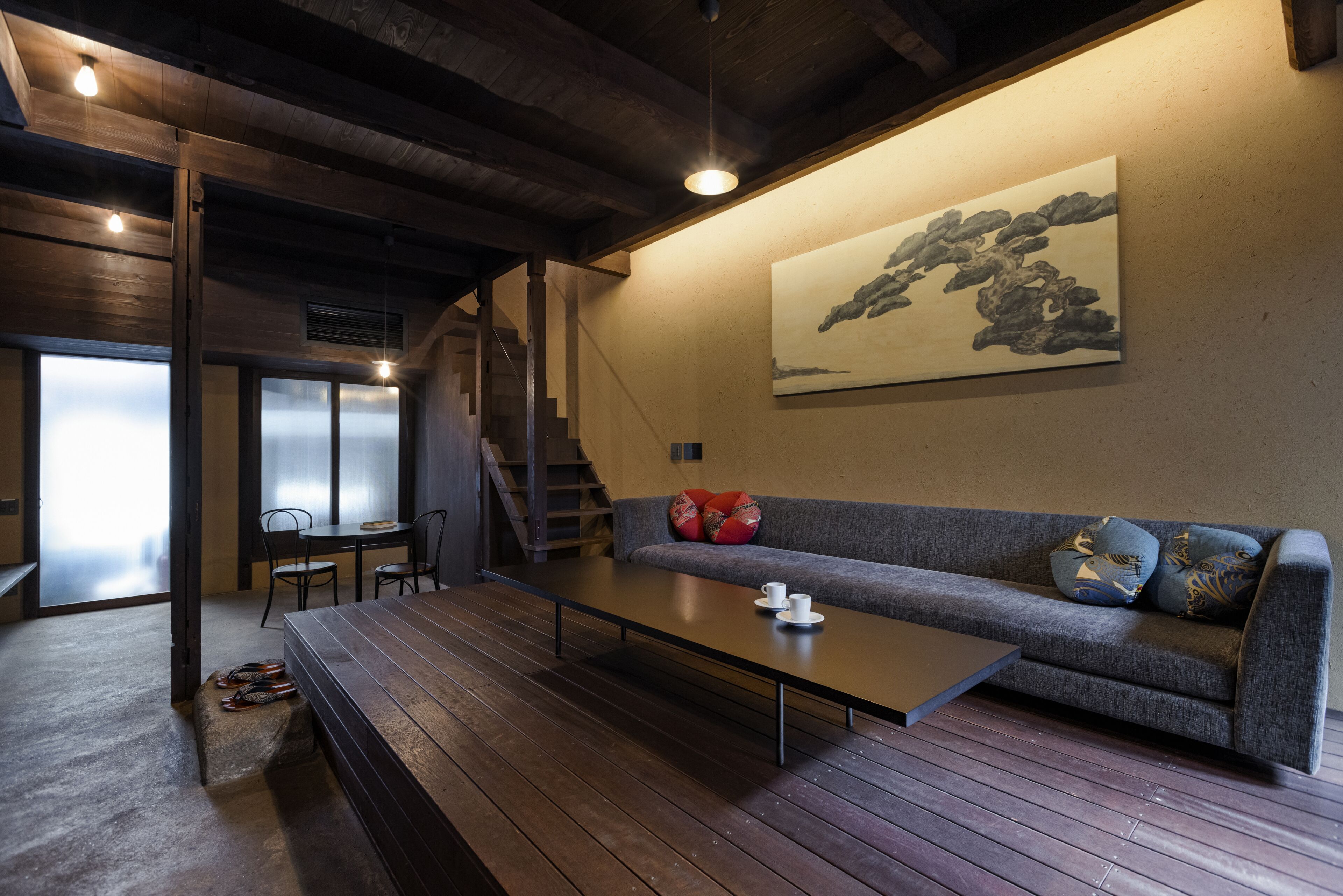 A modern living room with a wooden floor, a gray sofa with patterned cushions, a coffee table, and a staircase leading to another room.