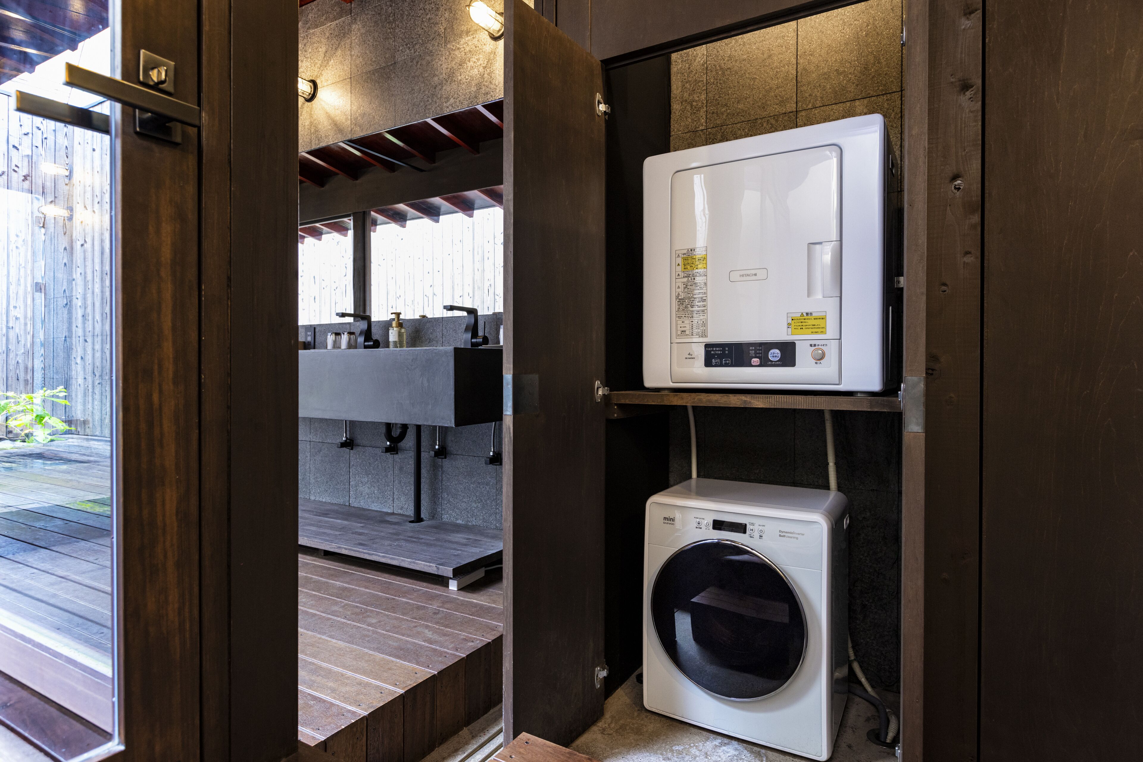 A compact laundry area with a front-loading washer and dryer, a wooden deck outside, and a modern kitchen visible through an open door.