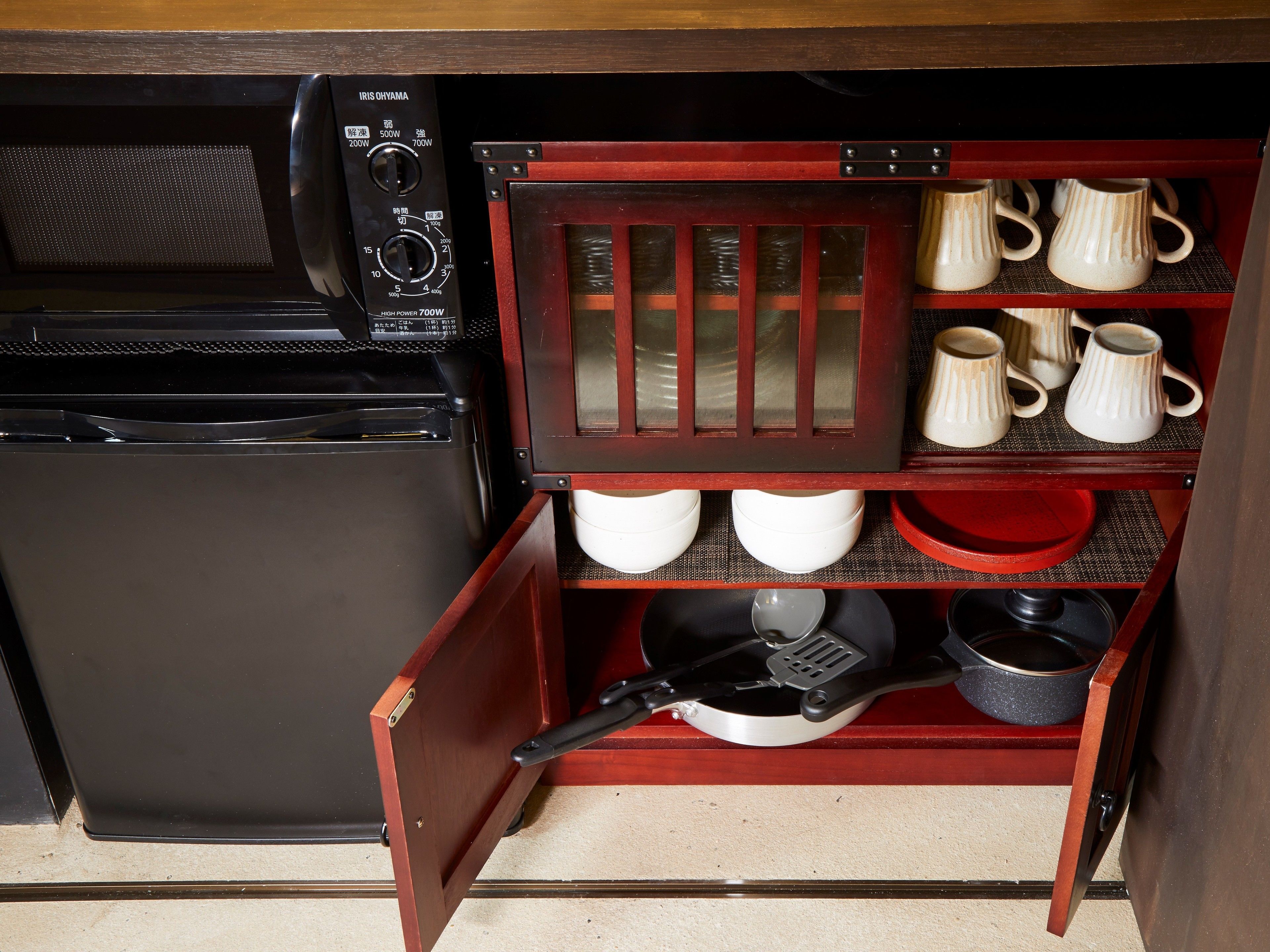 A compact kitchen with a microwave, a stove, and a wooden cabinet with shelves holding mugs, bowls, and a frying pan.