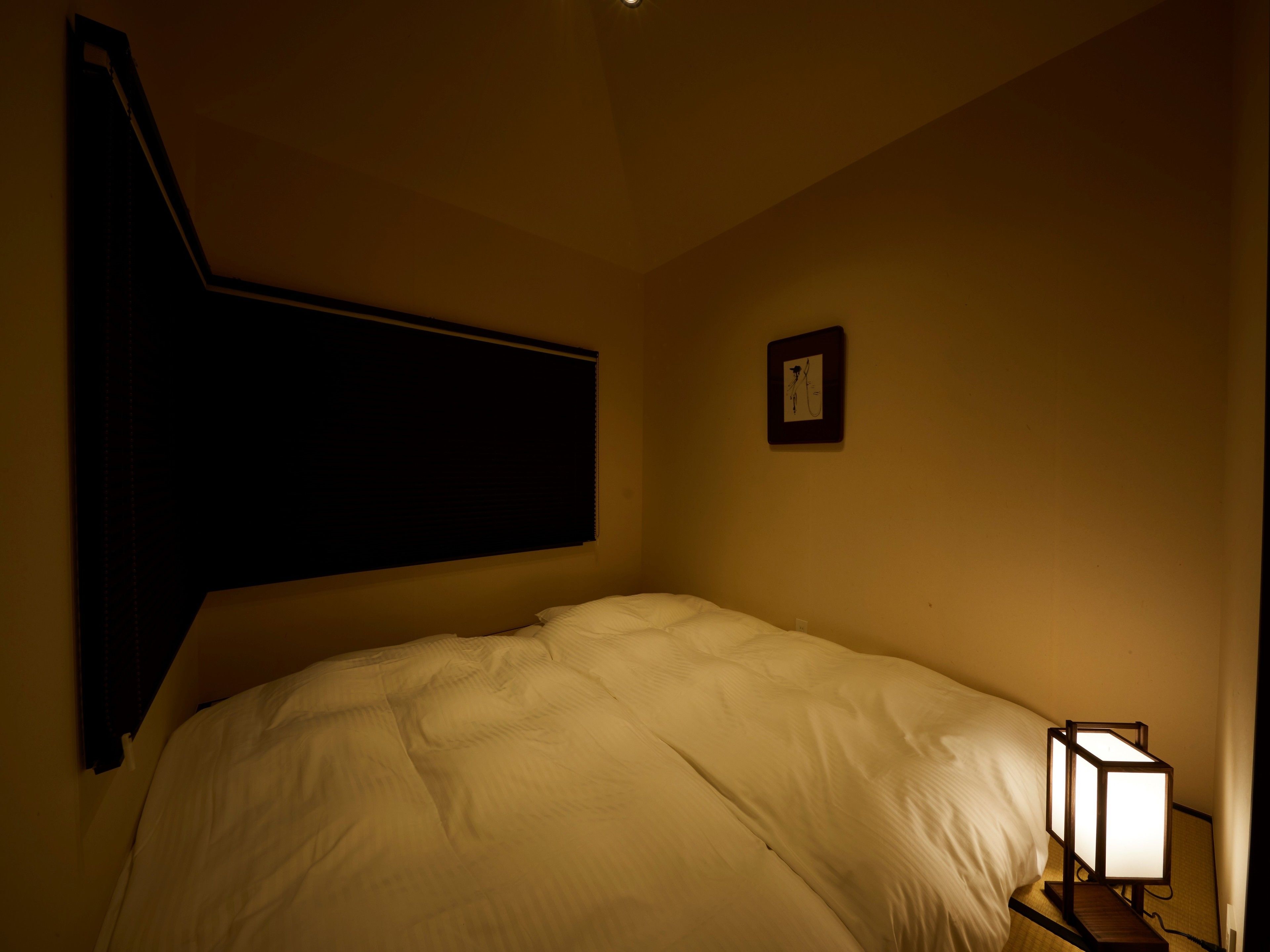 Habitación de hotel con una cama, una mesita de noche, un cuadro colgado en la pared y una ventana con persianas.