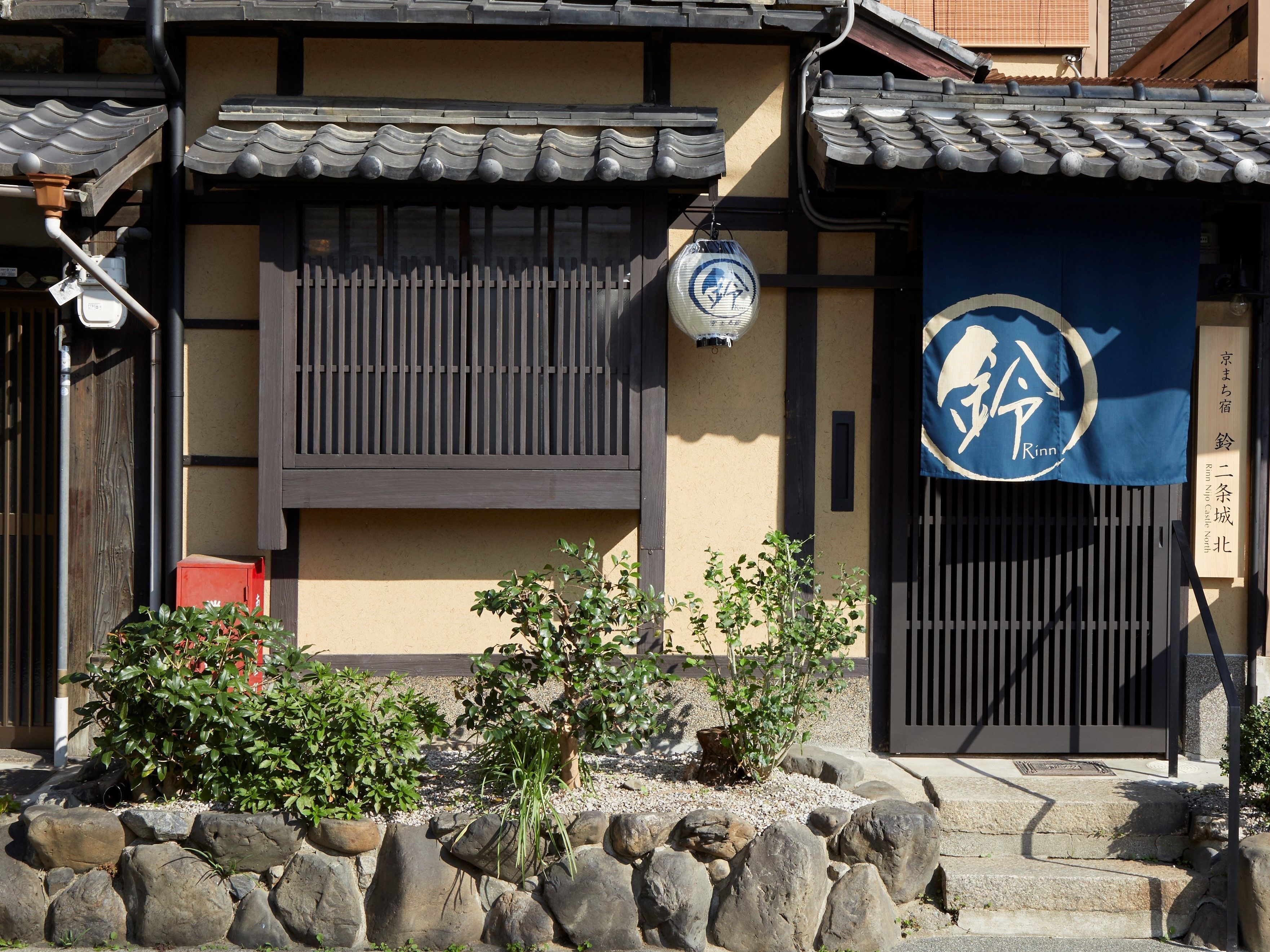 Un edificio japonés tradicional con techo de tejas, un banderín azul con texto blanco y una pared de piedra con plantas.