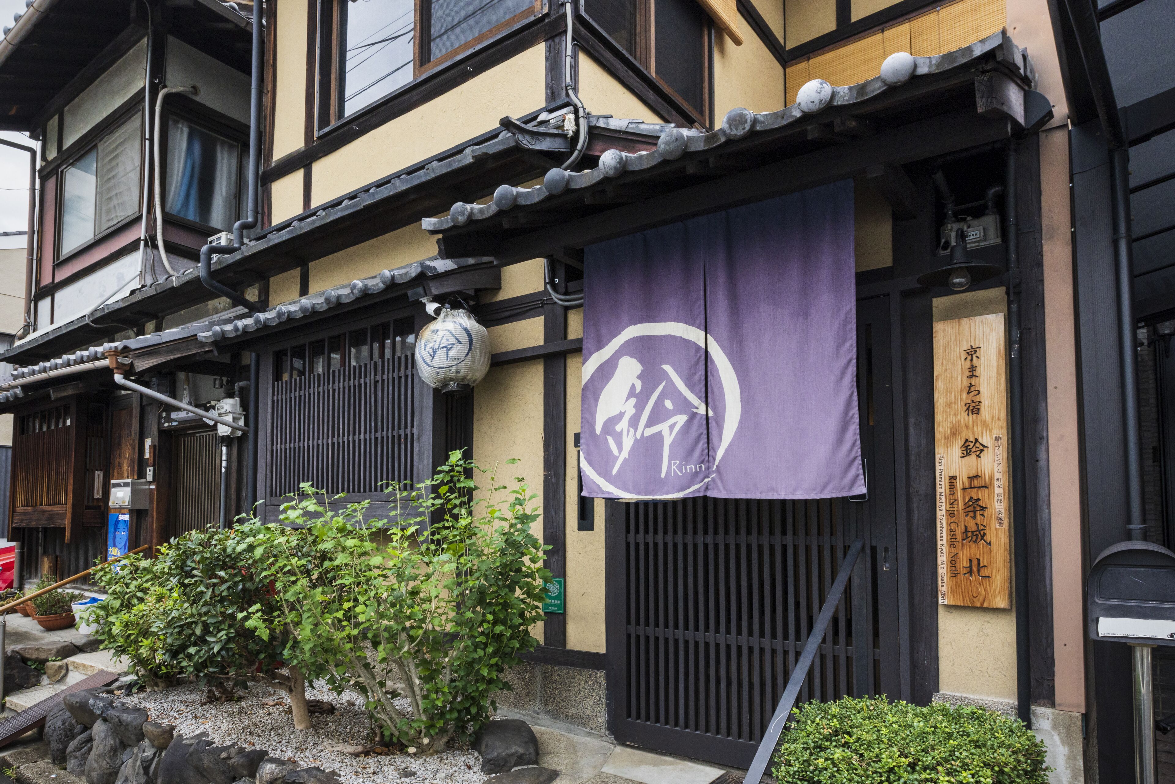 Un edificio japonés tradicional con un letrero de madera, un banderín morado con un logotipo y un pequeño jardín con rocas y plantas.