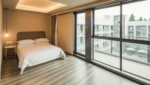 Ein modernes Hotelzimmer mit einem großen Bett, einem Nachttisch, einer Lampe und Blick auf die Stadt durch große Fenster.