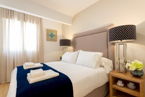 A neatly made bed with white linens and a blue accent, a bedside table with a lamp and a vase of flowers, and a window with curtains.