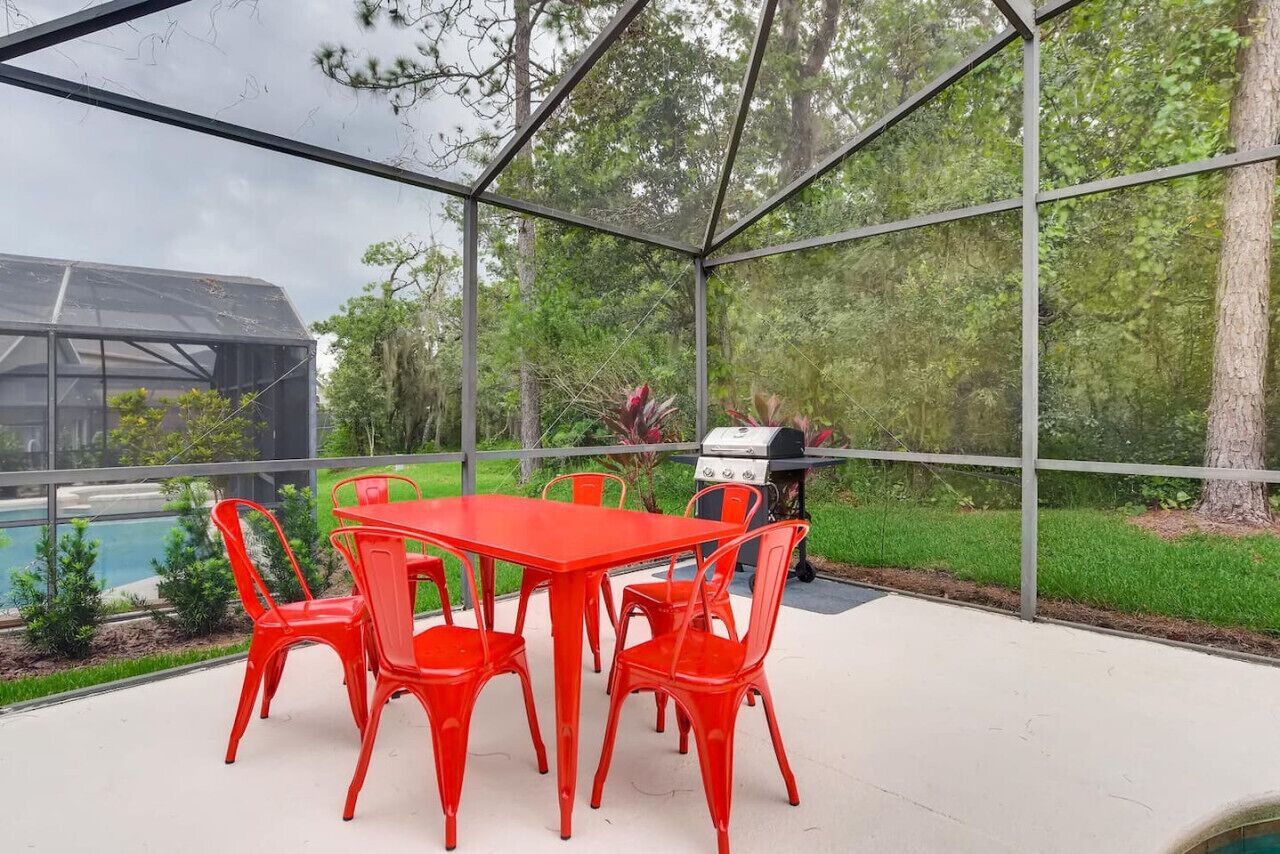 Un patio techado con una mesa y sillas rojas, una parrilla y vistas a una piscina y follaje.