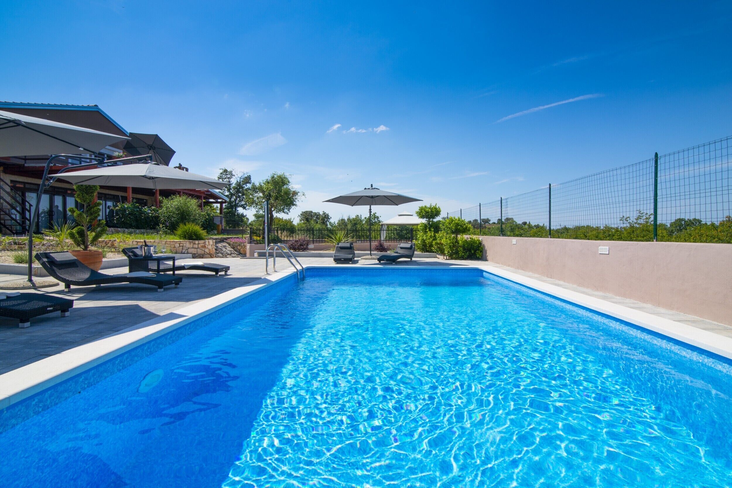 A swimming pool with lounge chairs and umbrellas, surrounded by a fence and greenery.