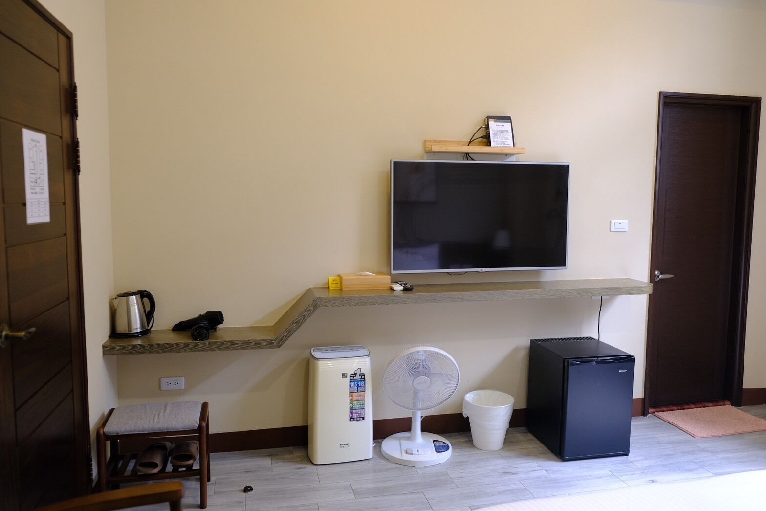 Una habitación con una televisión de pantalla plana montada en un estante de madera, un ventilador blanco, un refrigerador negro pequeño y una tetera en la encimera.