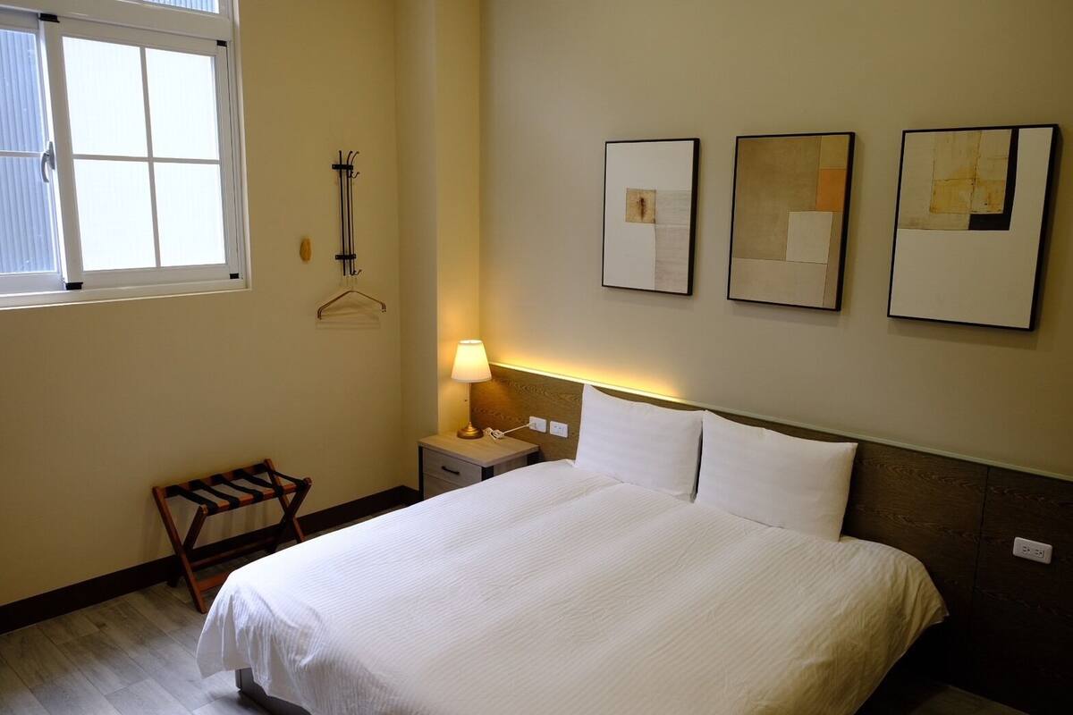 A hotel room with a bed, a nightstand with a lamp, a wooden bench, and three framed artworks on the wall.