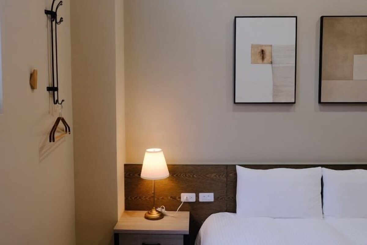 A hotel room with a bed, a nightstand with a lamp, and two framed artworks on the wall.