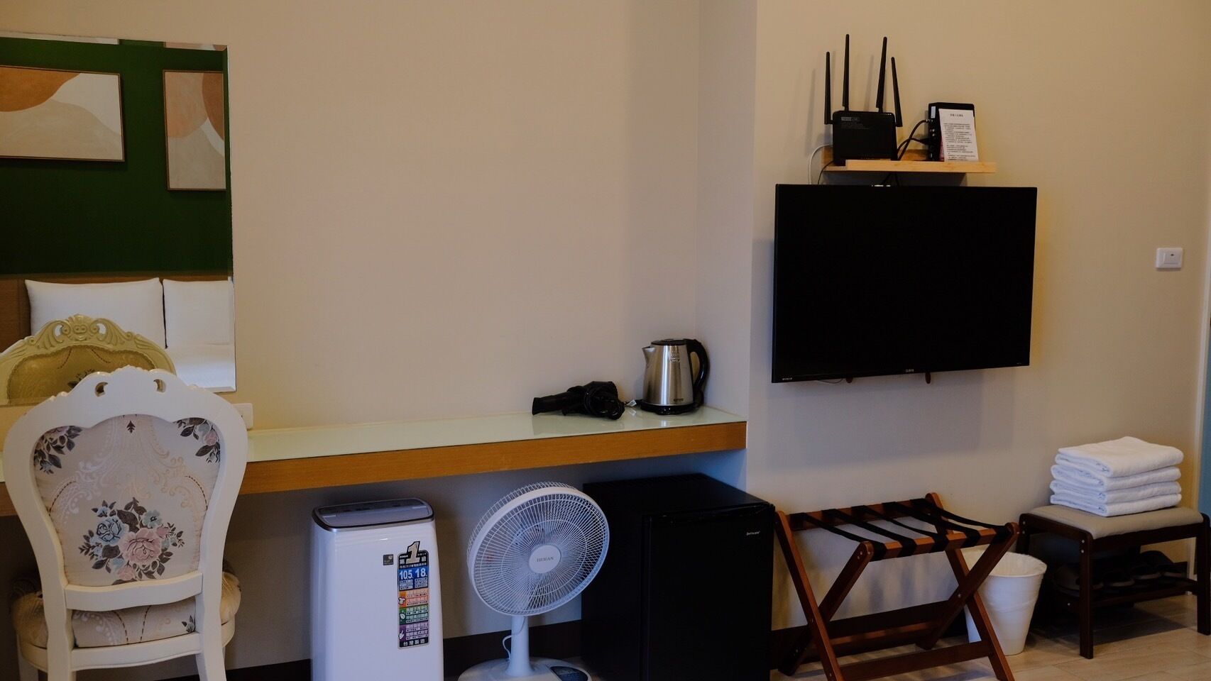 Et lille hotelværelse med seng, skrivebord med elkedel, ventilator, fjernsyn og stol.