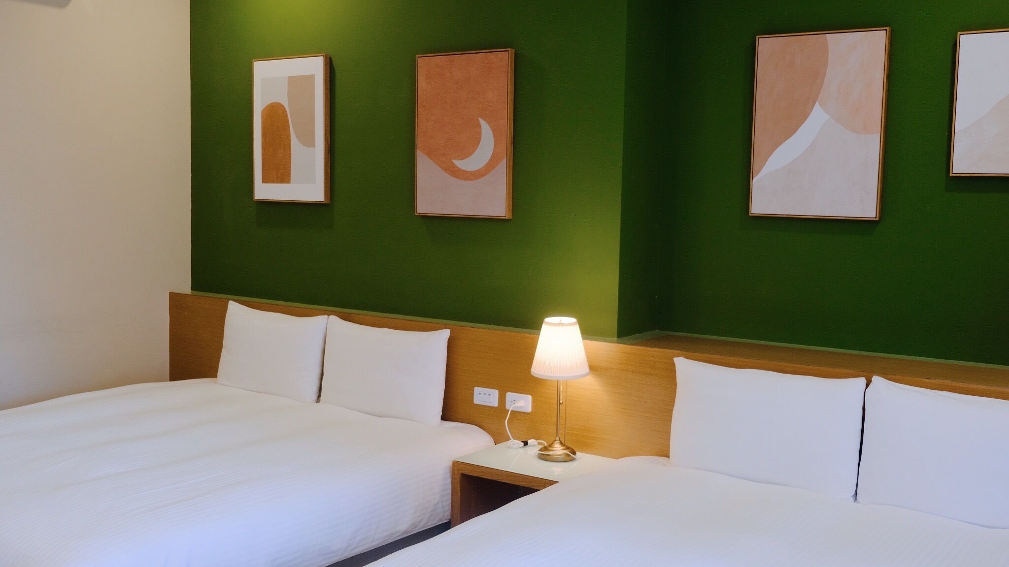A hotel room with two beds, a nightstand with a lamp, and abstract artwork on the green wall.