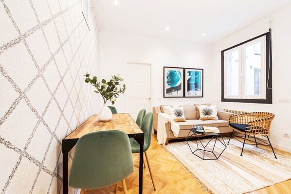 Ein modernes Wohnzimmer mit einem Holzesstisch, grünen Stühlen, einer Couch und einem Rattan-Sessel. An der Wand hängen zwei gerahmte Kunstwerke und ein Fenster mit Jalousien.