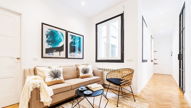 A modern living room with a sofa, a coffee table, and a wicker chair. There are two framed artworks on the wall and a window with blinds.
