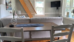 Un comedor con una mesa de madera, sillas blancas, una escalera de madera, un televisor montado en la pared y un ventanal con vistas.