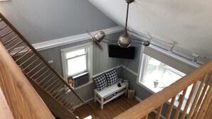 Una habitación con techos altos, una escalera de madera, un ventilador de techo, un televisor montado y una ventana con persianas.