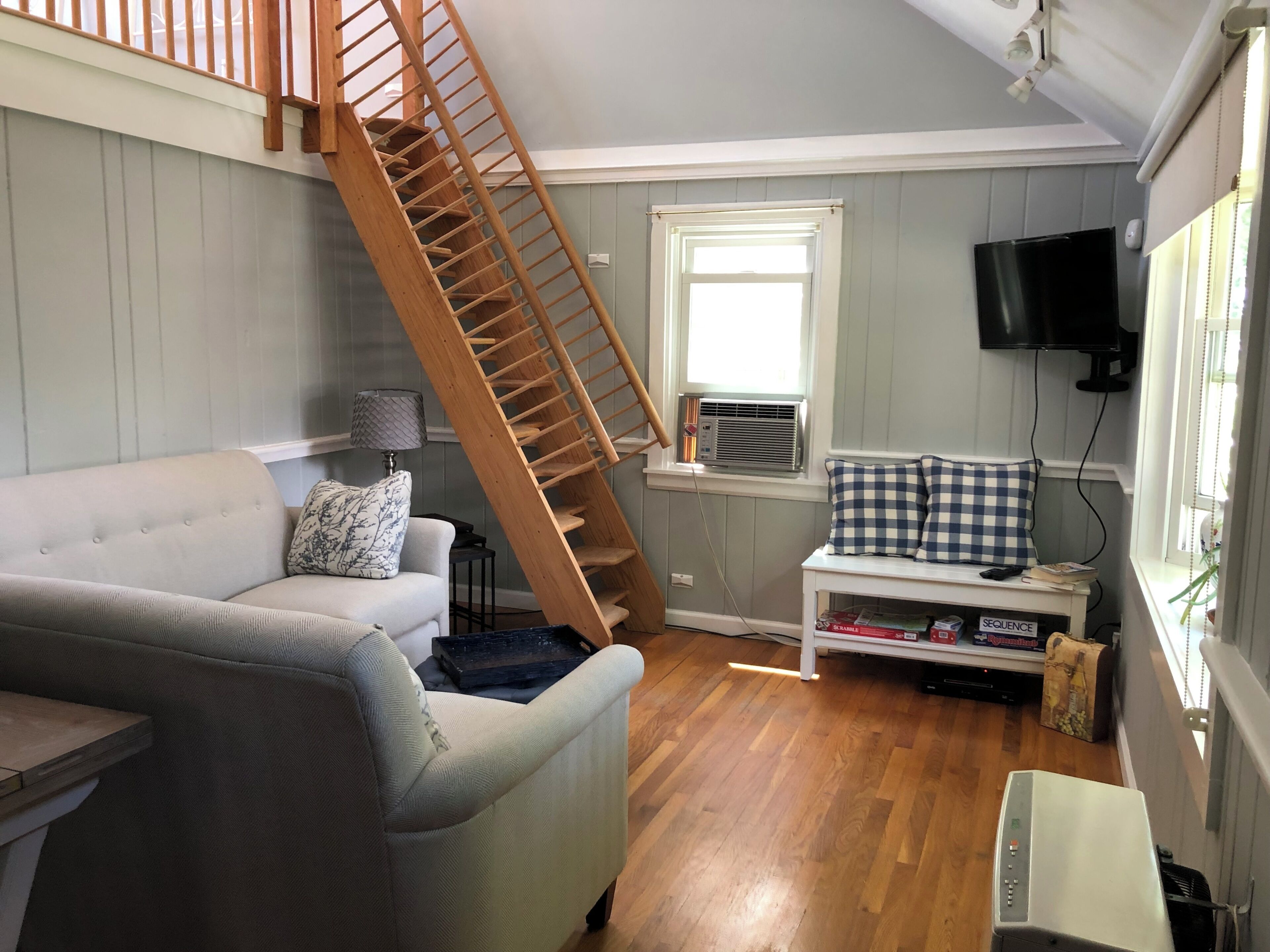 A living room with a wooden staircase, a grey sofa, a small table with a lamp, a window with an air conditioner, and a television mounted on the wall.