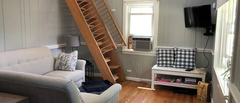 A living room with a wooden staircase, a grey sofa, a small table with a lamp, a window with an air conditioner, and a television mounted on the wall.