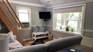 Una sala de estar acogedora con un sofá gris, una mesa de madera, un televisor montado en la pared y un ventanal con vistas a la vegetación.