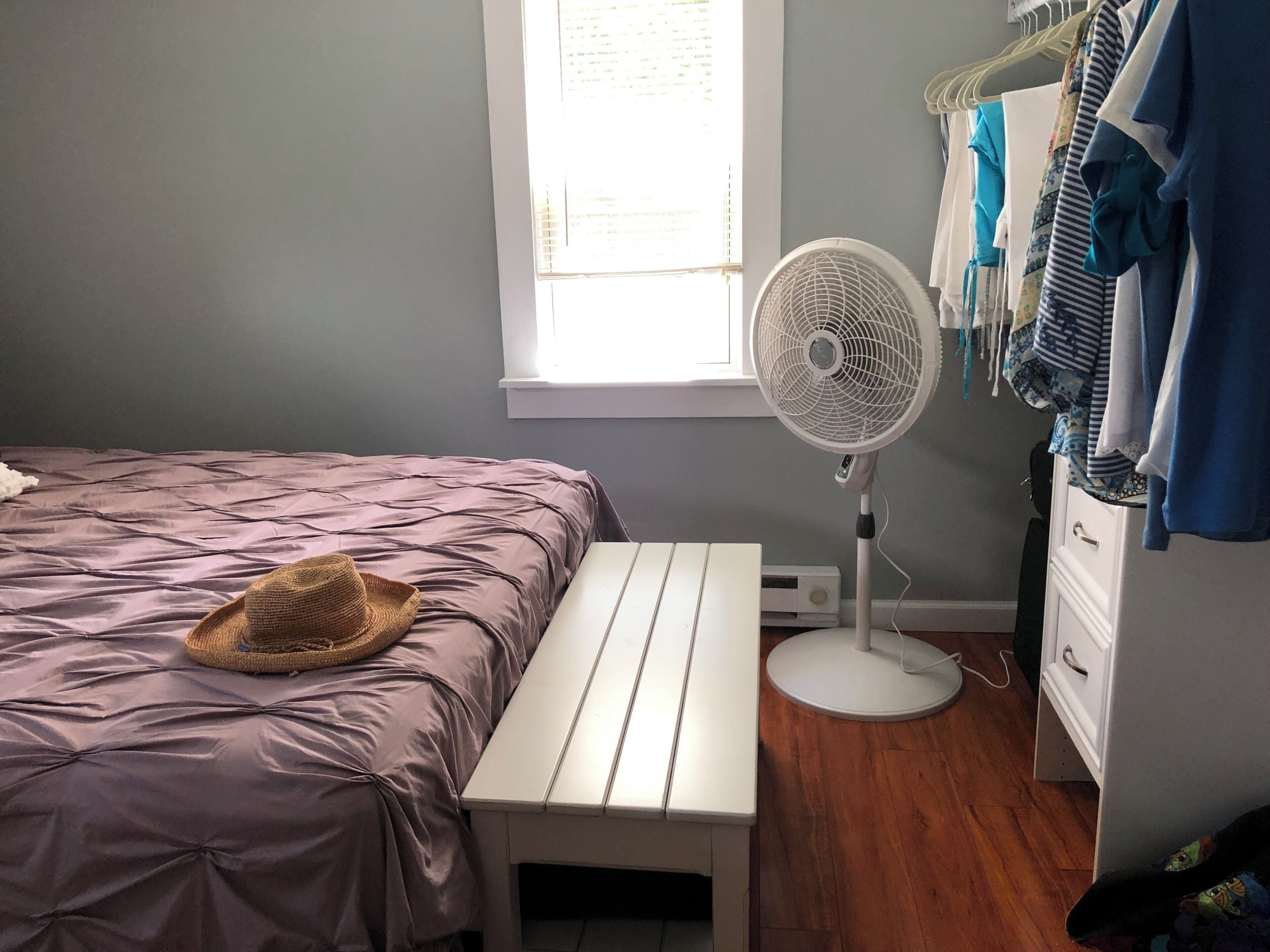Une chambre à coucher comprenant un lit, une table de chevet, un ventilateur, une fenêtre et une armoire.