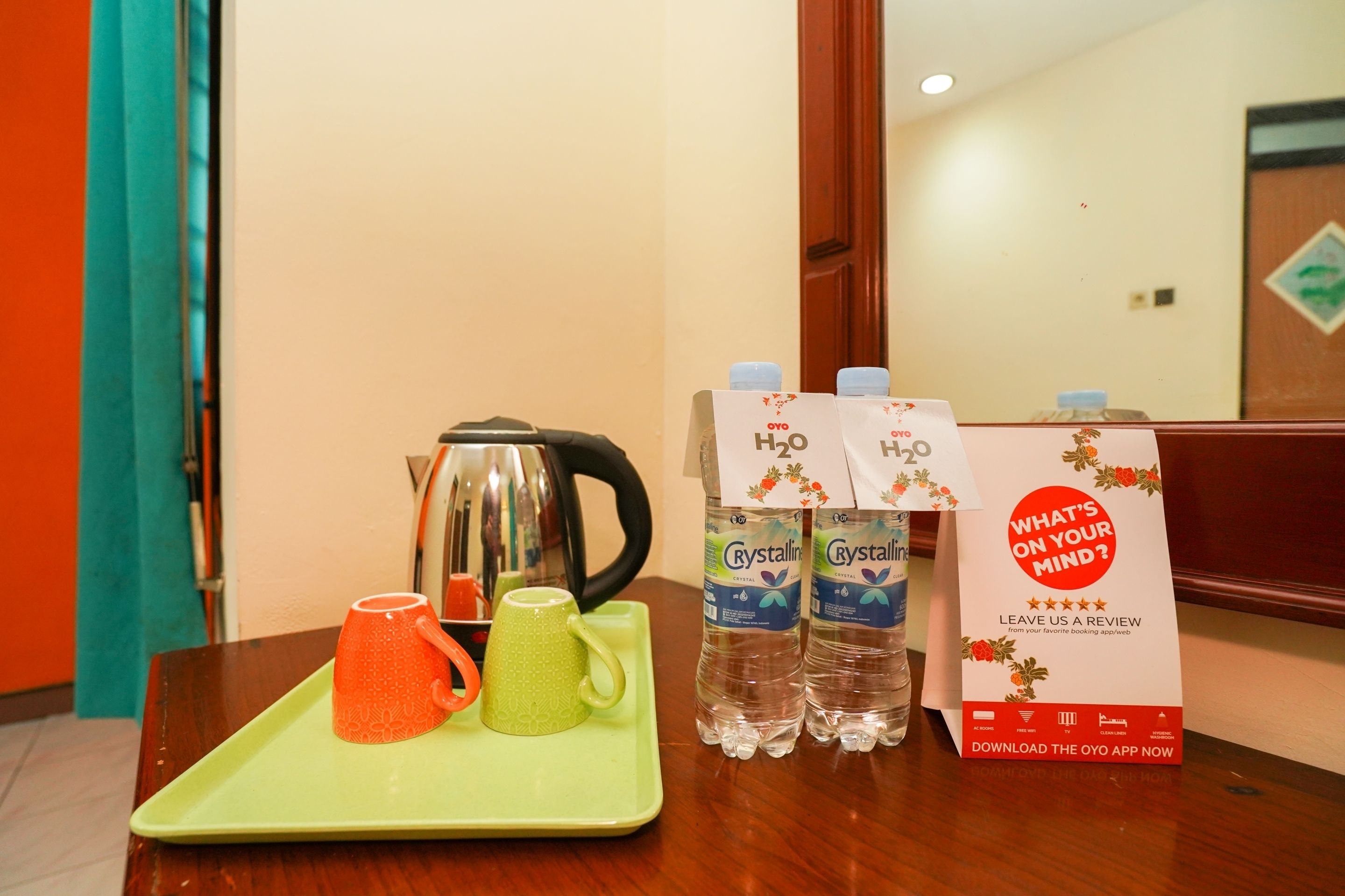 A table with a tray holding two mugs, a kettle, and two bottles of water. There is a carton of OYO branded water and a sign asking for a review.