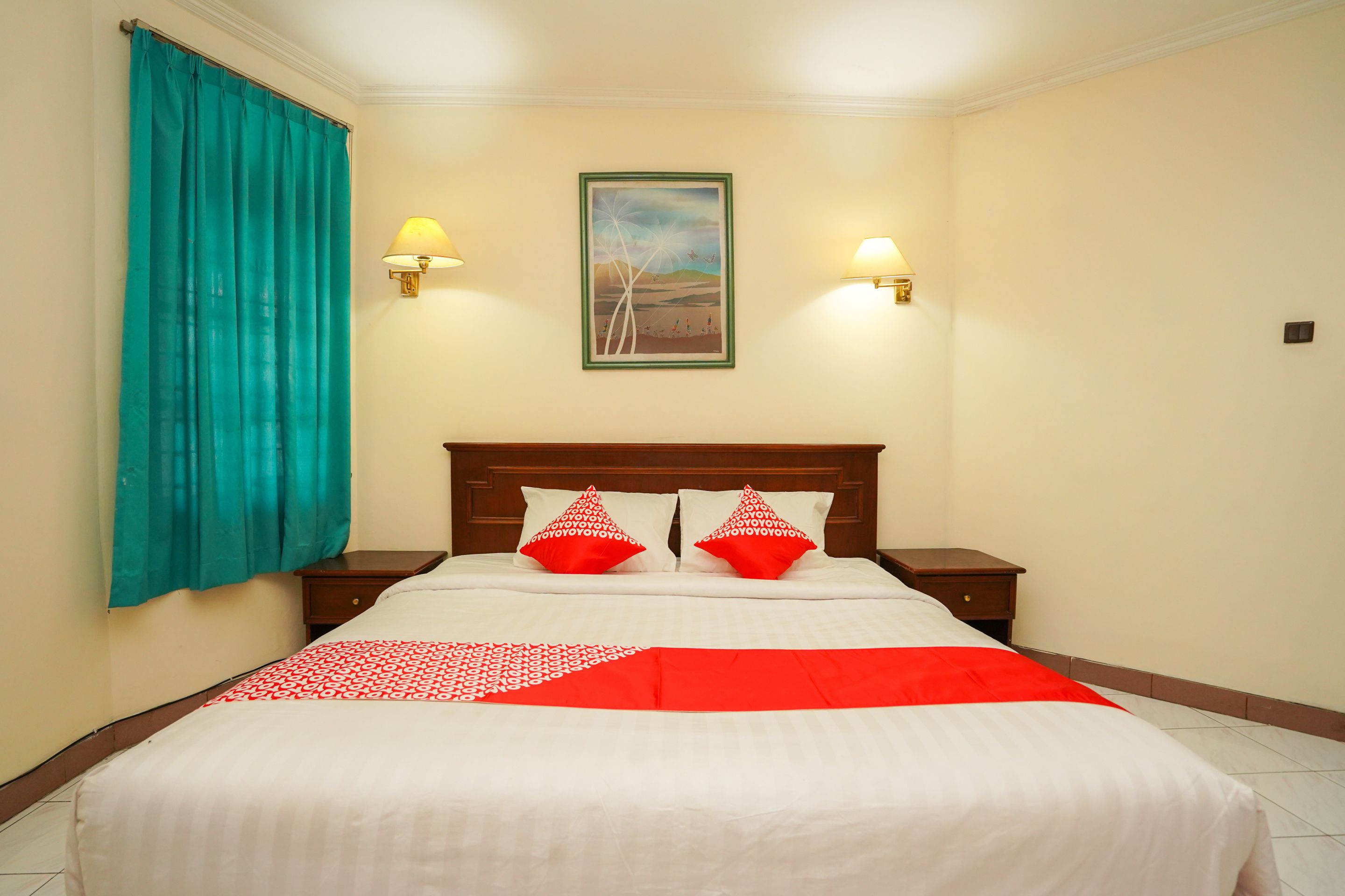 Une chambre d’hôtel avec un lit, des rideaux turquoise, deux lampes de chevet, un tableau encadré au mur et deux coussins rouges.