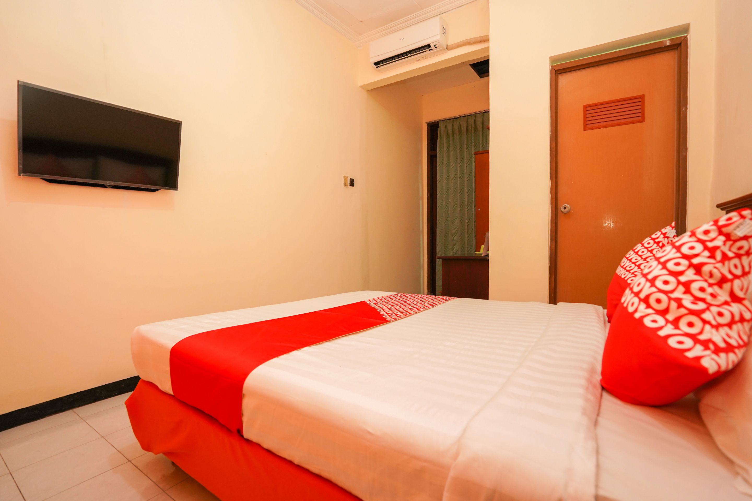A hotel room with a bed, a flat-screen TV mounted on the wall, and an air conditioning unit above the door.