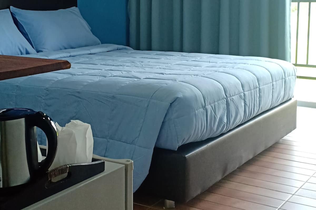 A bedroom with a bed, a bedside table with a kettle, and a window with curtains.