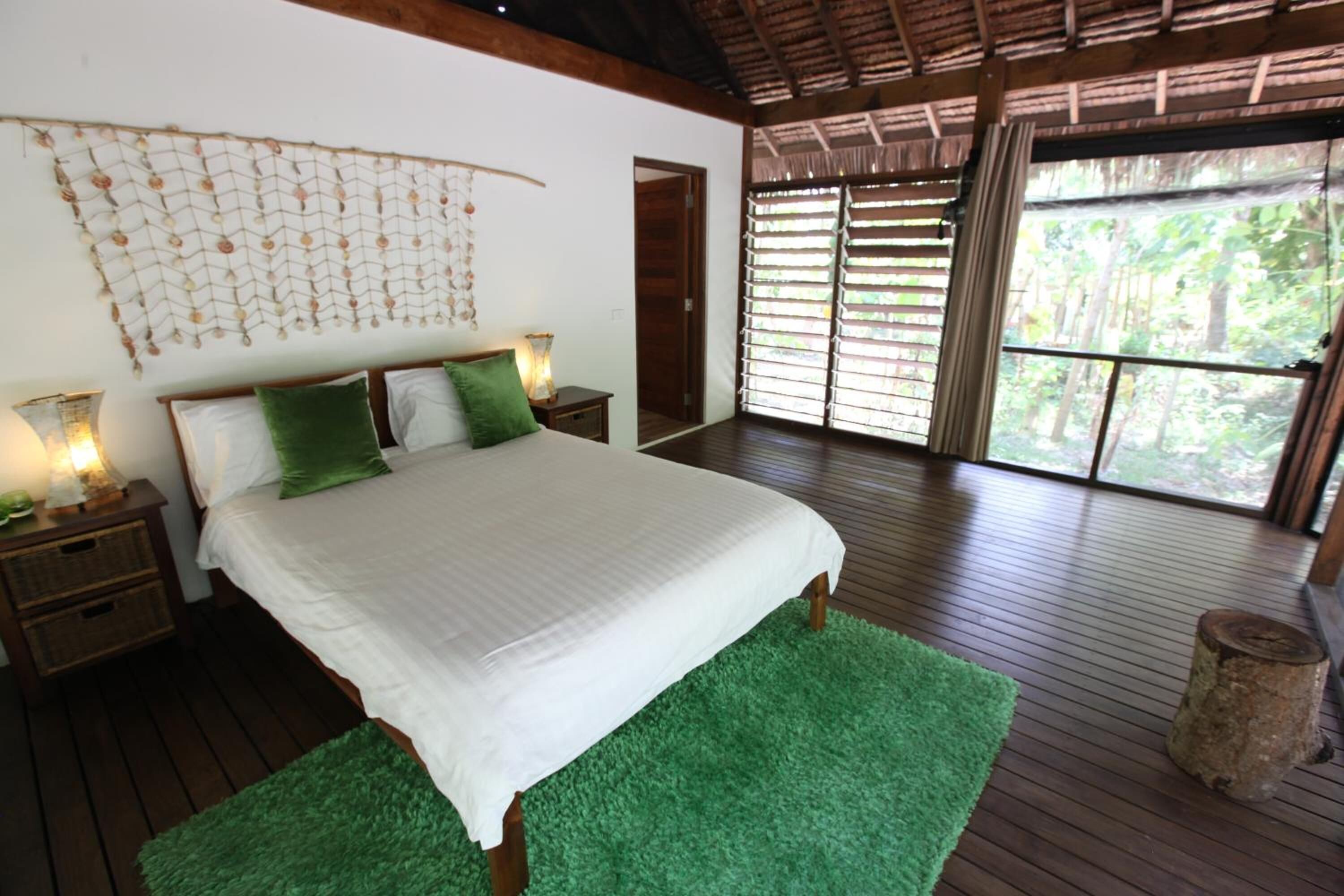 A bedroom with a large bed, wooden floors, a green rug, a wall hanging, and a window with blinds.