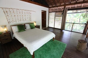 A bedroom with a large bed, wooden floors, a green rug, a wall hanging, and a window with blinds. - Divi's Boutique Retreat (Mangaliliu)