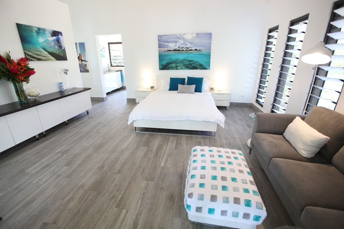 A modern hotel room with a large bed, a sofa, a small table, and a view of the ocean through the windows.