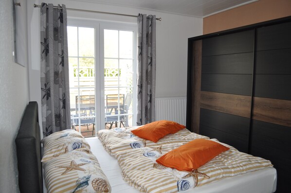 A double bed with striped bedding and two orange pillows, a dark wooden headboard, and a view of an outdoor patio with chairs and a table. - Holiday home in a quiet location in the center of Nordseebad Tossens (Tossens)