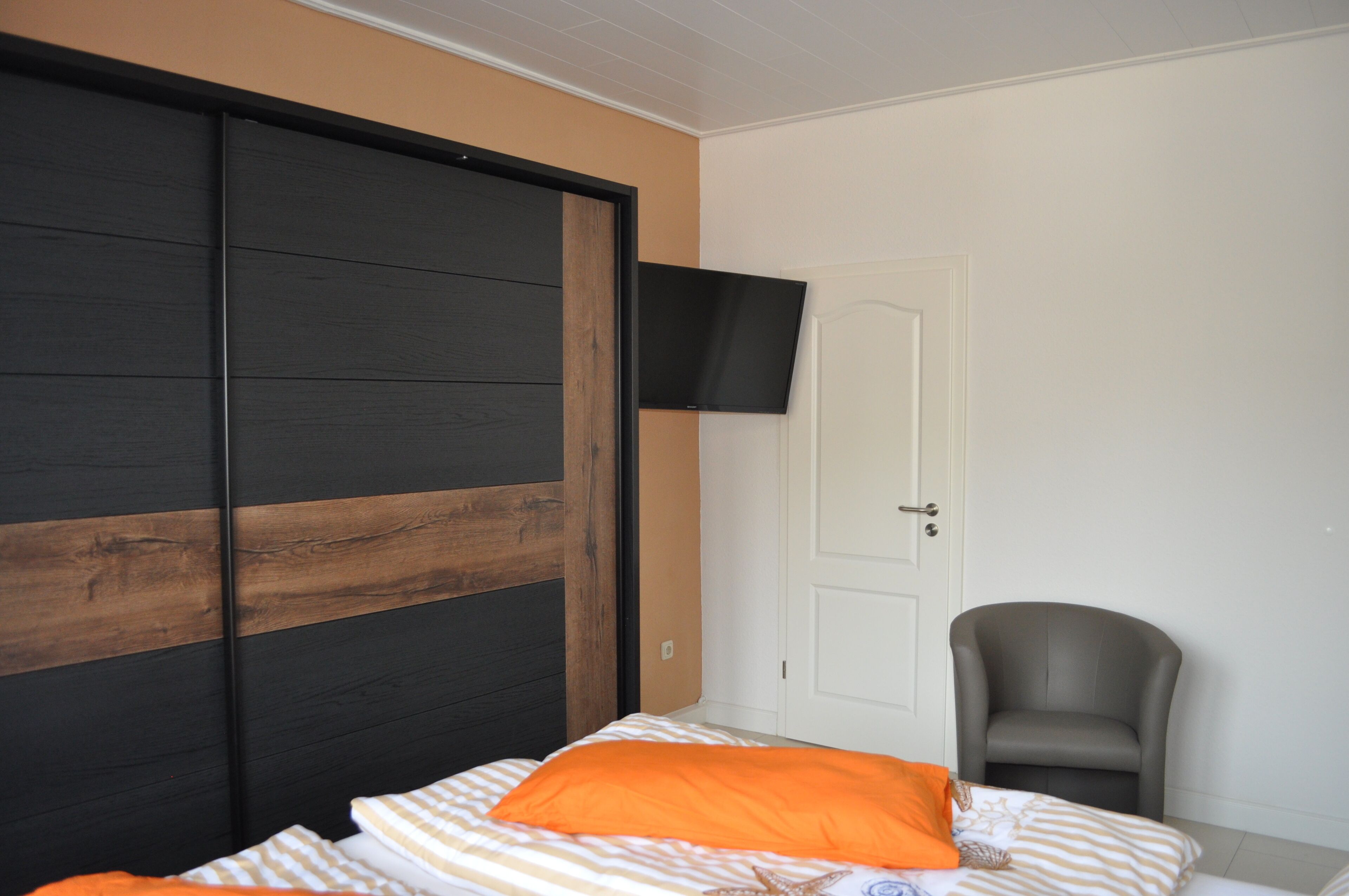 A bedroom with a bed, an orange pillow, a grey chair, a TV mounted on the wall, and a door.