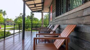 Een overdekt terras met houten banken en tafels, een metalen hekwerk en uitzicht op de omliggende begroeiing en de weg.