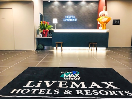 A reception area with a black reception desk, two wooden stools, a potted plant, and a sign reading "HOTEL LIVEMAX".