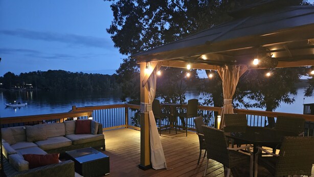 A wooden deck with outdoor furniture, string lights, and a view of a lake at dusk. - FREE USE OF OUR 2022 23’ TRITOON EVERY RENTAL DAY
May 1st- Oct 31st (Albemarle)