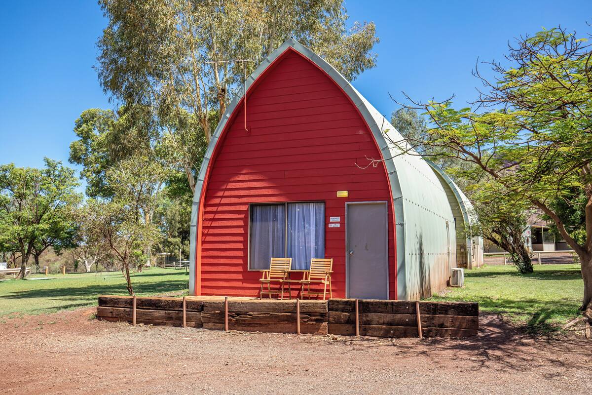 A red barn with a door and windows, wooden chairs outside, and trees in the background.