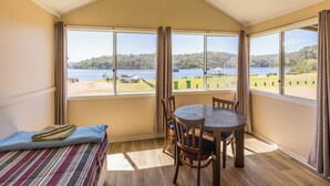 A room with a bed, a table, and chairs, large windows with curtains, and a view of a lake and greenery. - Walpole Rest Point Caravan Park (Walpole)