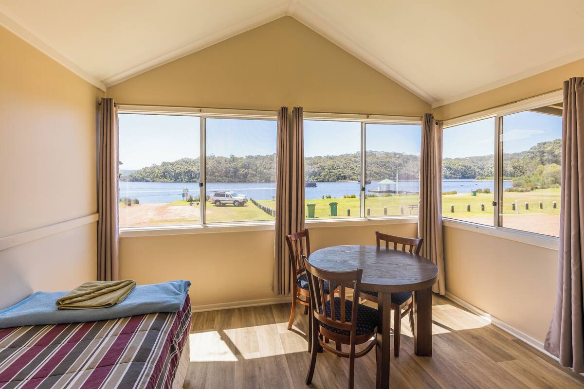 A room with a bed, a table, and chairs, large windows with curtains, and a view of a lake and greenery.