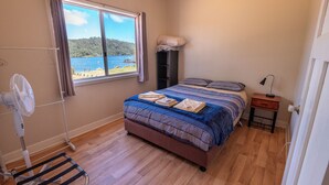 A bedroom with a bed, a fan, a window with a view of the sea, and a small bedside table. - Walpole Rest Point Caravan Park (Walpole)