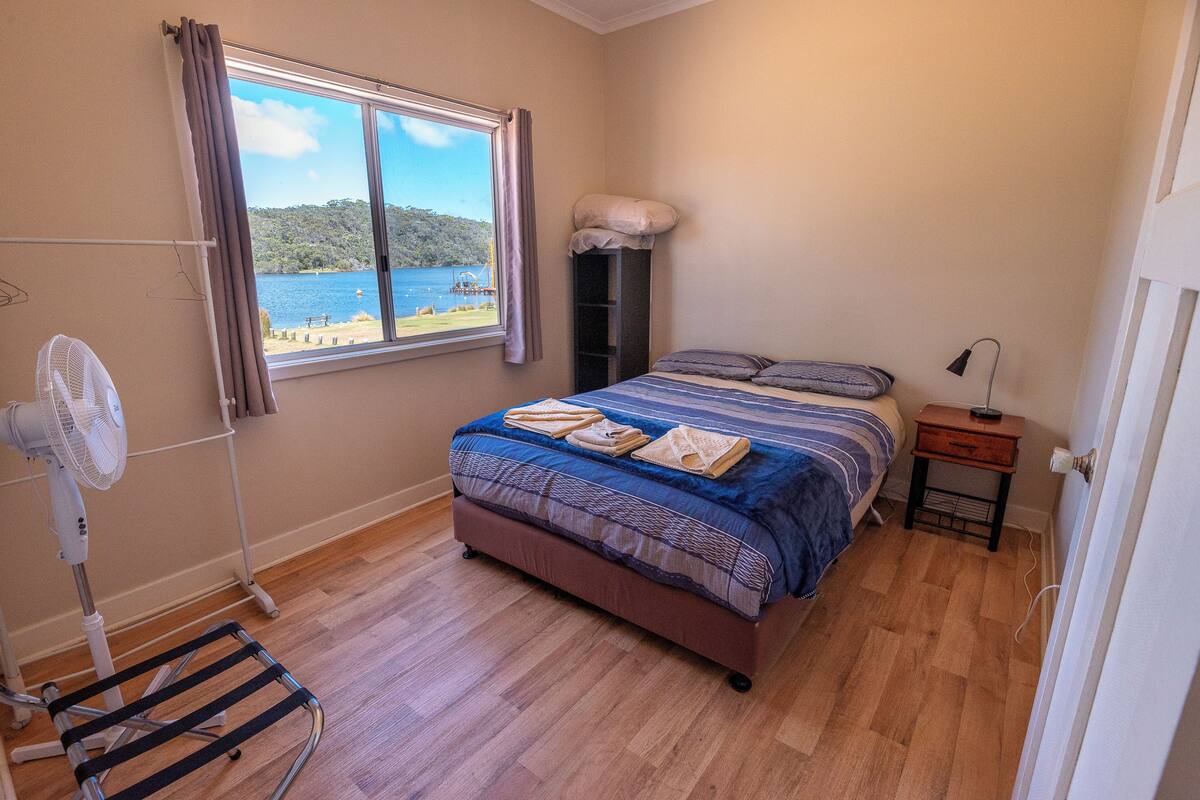 A bedroom with a bed, a fan, a window with a view of the sea, and a small bedside table.