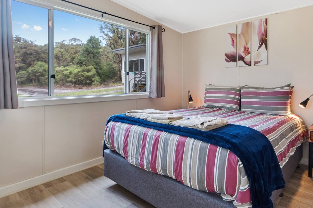 A bedroom with a large window, a bed with striped bedding, a bedside table, and a wall-mounted lamp.