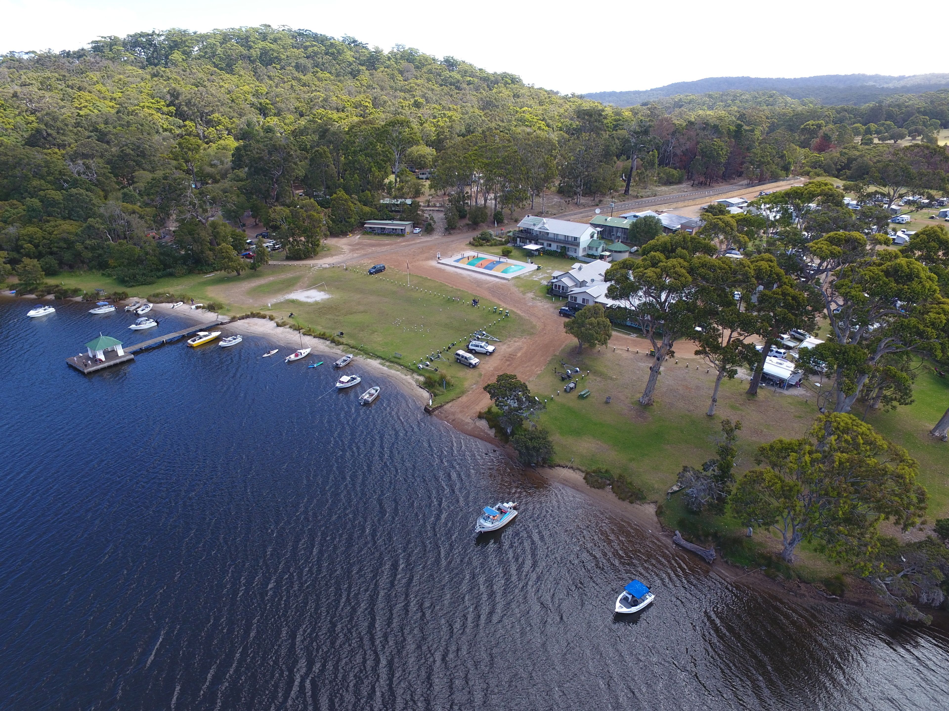 An aerial view of a lakeside property with a dock, boats, and a building with a swimming pool.