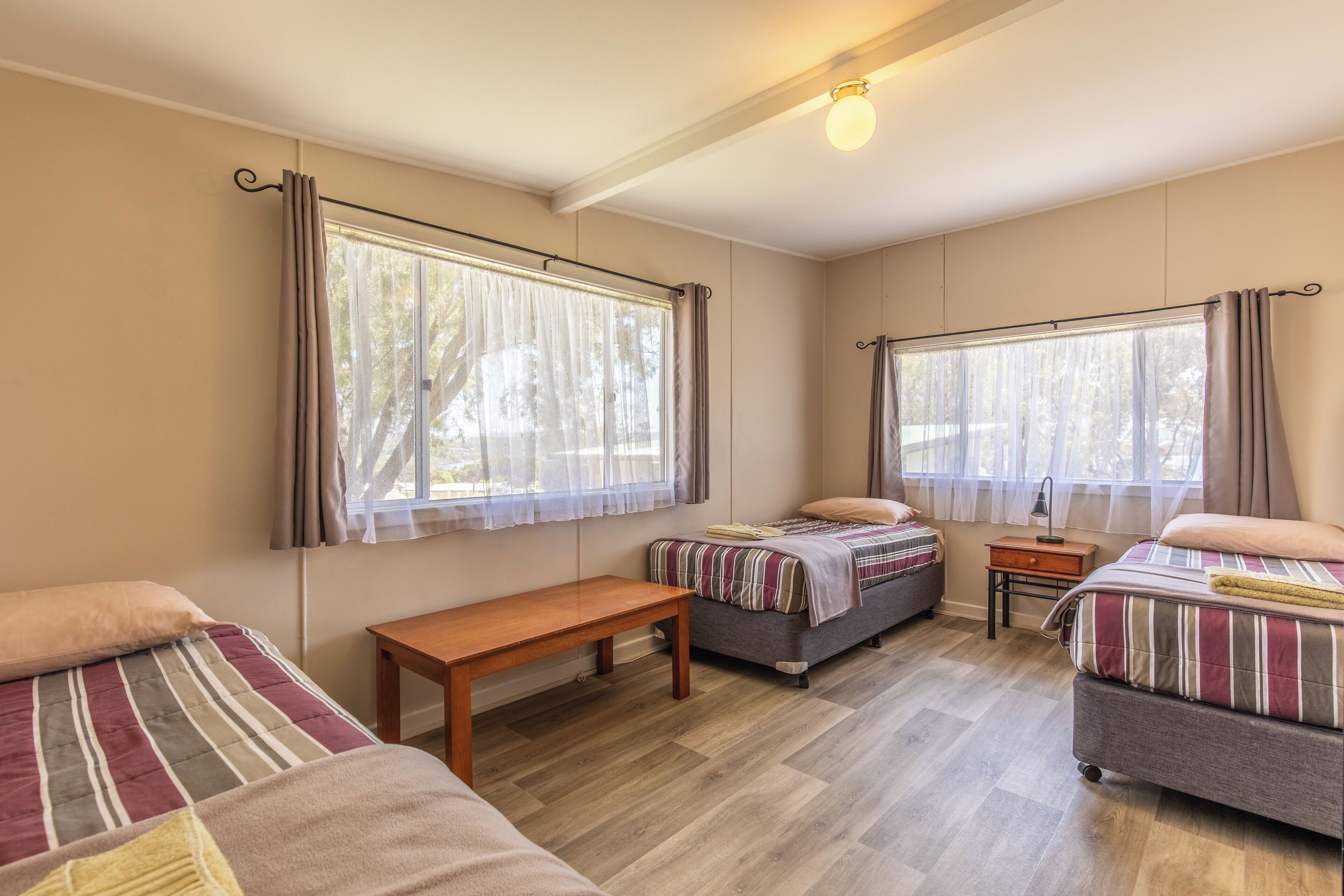 A room with two beds, a bench, a nightstand, and large windows with curtains.