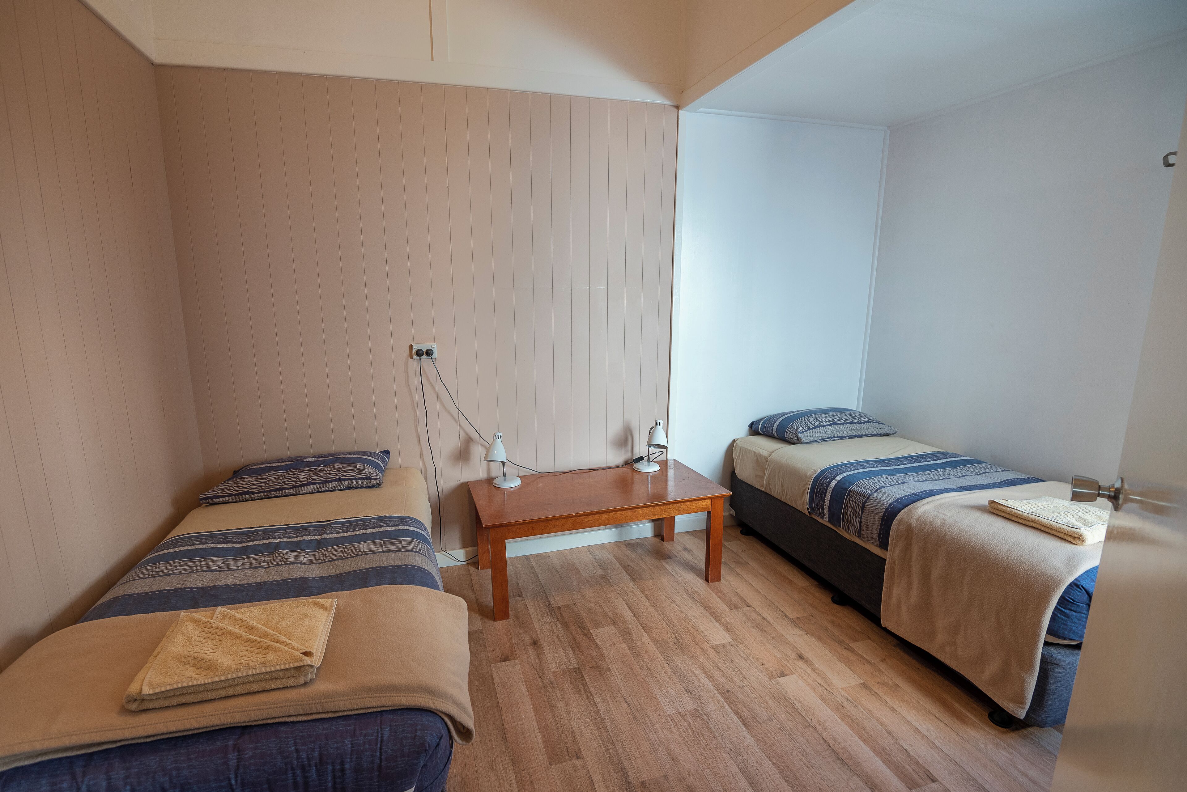 A small, well-lit room with two single beds, a wooden table, and a white wall.