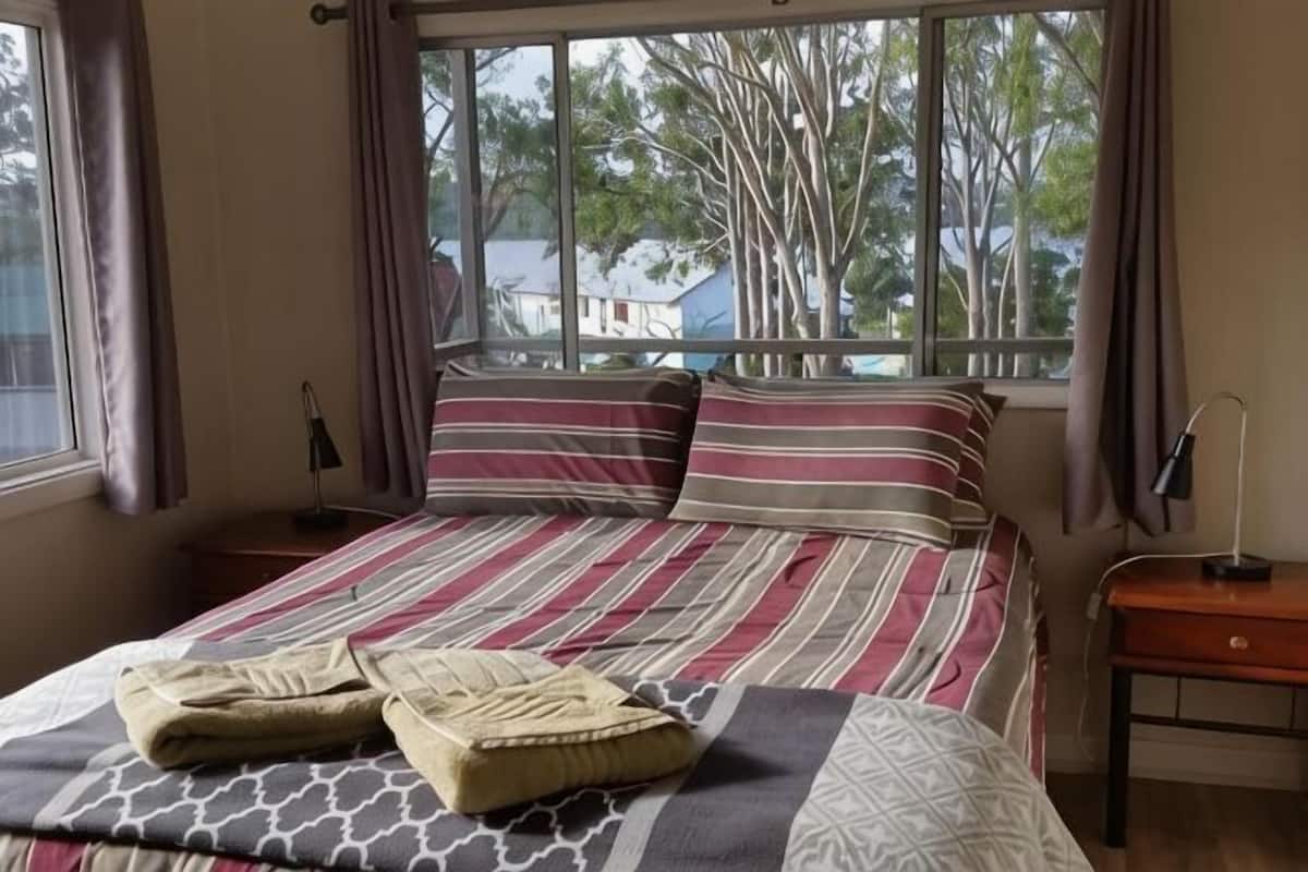 Una cama con ropa de cama a rayas, una ventana con vista a árboles y una mesita de noche con una lámpara.