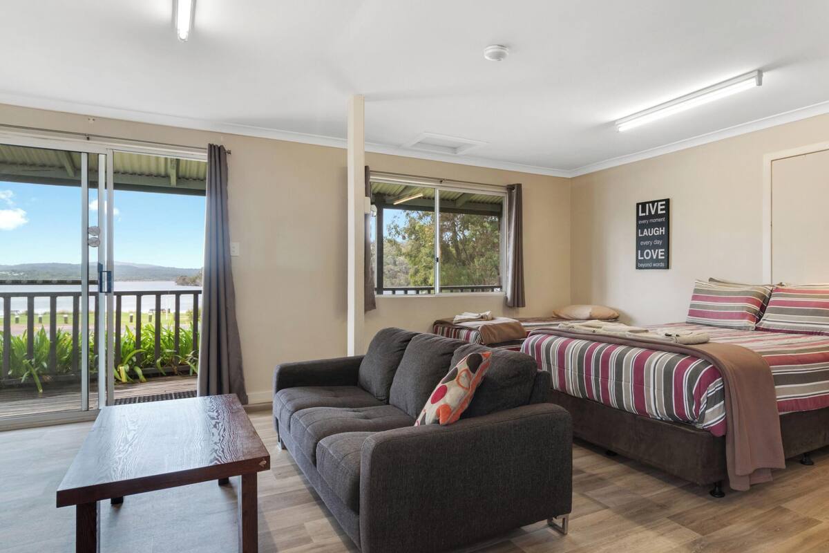 A room with a sofa, a bed with striped bedding, a table, and a view of the outdoors through a sliding door.