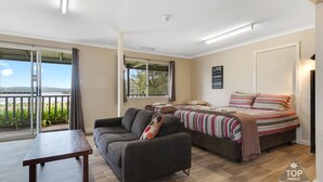 A modern bedroom with a bed, sofa, and a table. There is a balcony with a view of greenery and a clear sky. - Walpole Rest Point Caravan Park (Walpole)