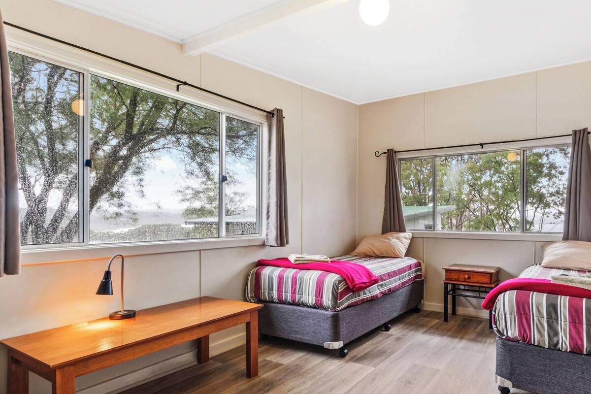 A room with two beds, a wooden bench, a small table, and large windows with curtains.